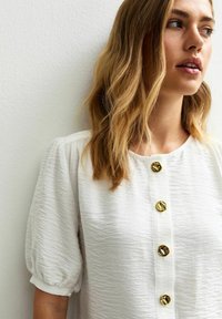 Witte textuur blouse met korte pofmouwen, voorzien van vijf decoratieve gouden knopen aan de voorkant. Eenvoudig en casual ontwerp.