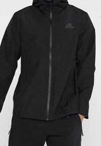 Svart lättviktsjacka med dragkedja, justerbar huva och Adidas-logotyp. Har en slät yta och smal passform, perfekt för lager-på-lager.