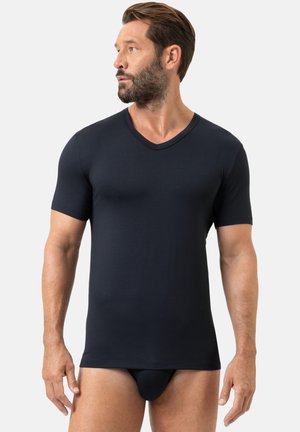 Mann mit Bart, der ein schwarzes, eng anliegendes T-Shirt mit kurzen Ärmeln und V-Ausschnitt sowie schwarze Slips trägt und vor einem hellen Hintergrund seitlich schaut.