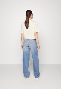 Žena s dlouhými hnědými vlasy v culíku, oblečená v béžovém tričku s krátkým rukávem a volných světle modrých džínách, stojí čelem k obyčejné bílé zdi.