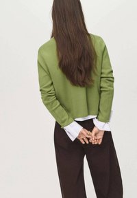 Pull vert court avec des manches longues, superposé à une chemise blanche ; le mannequin a de longs cheveux bruns et porte un pantalon marron foncé.