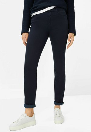 Marineblaue Slim-Fit-Hosen mit umgeschlagenen Säumen, aus einem weichen Baumwollgemisch. Kombiniert mit weißen Schnürsneakern vor einem schlichten Hintergrund.