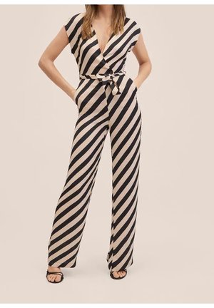 Kvinde iført en beige og sort diagonalt stribet ærmeløs jumpsuit med en dyb V-udskæring og taljebånd, der står med hænderne i lommerne.