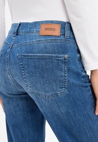 Gros plan sur une personne portant un jean en denim bleu avec une poche arrière et une étiquette en cuir portant le mot "ANGELS", associé à une chemise blanche à manches longues.