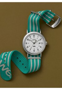Timex Weekender óra fehér számlappal, ezüst színű tokjával és türkiz csíkos szövet szíjával. Könnyen olvasható számokkal és dátumfunkcióval rendelkezik.