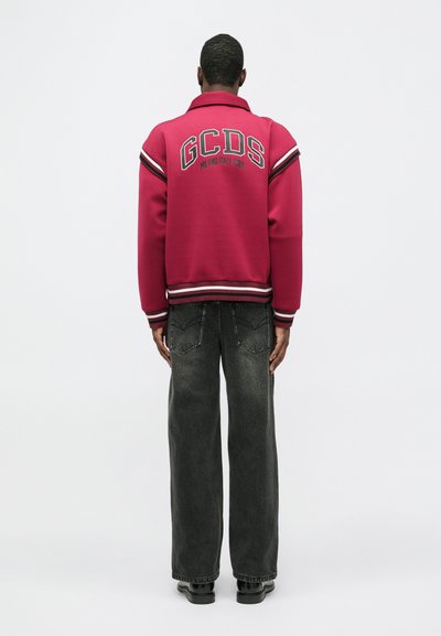 Moški v rdeči jakni varsity z napisom GCDS Milano Italija 1988 na hrbtni strani, v kombinaciji s širokimi črnimi jeans hlačami in črnimi čevlji, stoji obrnjeno stran od kamere.