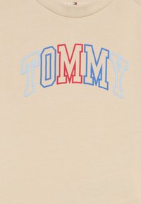 Μπεζ βαμβακερό T-shirt με τη λέξη "TOMMY" σε έντονα, καμπυλωτά γράμματα, με κόκκινες και μπλε λεπτομέρειες. Μαλακή υφή, στρογγυλή λαιμόκοψη.