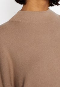Pull marron à côtes avec un design à col montant. Le tissu est lisse avec un léger éclat, créant une texture douce visuellement attrayante.