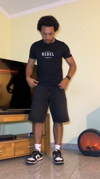 Schwarzes T-Shirt mit dem Aufdruck "REBEL", Jeansshorts, schwarze Sneaker mit weißen Akzenten, stehend drinnen mit einem Fernseher und einem dekorativen Heizkörper.