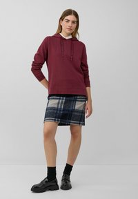 Sudadera con capucha color marrón burdeos y cordones, combinada con una falda de lana a cuadros en tonos navy y crema. Unos mocasines de charol negros y calcetines negros completan el look.