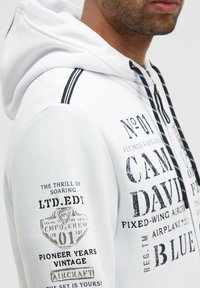 Sudadera blanca con capucha, con estampado de texto negro y diseños gráficos. Cuenta con cordones, franjas laterales y una tela suave y texturizada.
