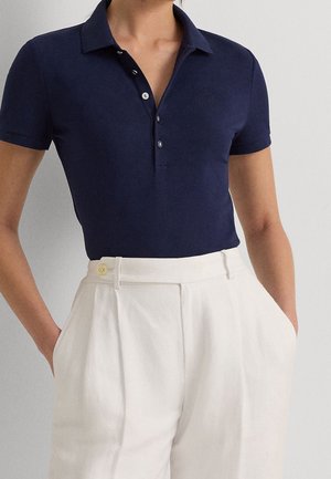 Personne portant un polo bleu marine à manches courtes rentré dans un pantalon blanc plissé taille haute, les mains dans les poches.