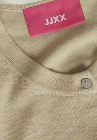 Beige neulottu neulepaita, jossa on pyöreä pääntie, nappikiinnitys ja vaaleanpunainen etiketti, jossa on valkoiset tekstit "JJXX." Kankaan rakenne on teksturoitu.