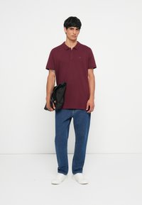 Burgunderfarget polo-skjorte med krage, korte ermer og en liten logo. Båret med blå jeans og hvite sneakers, holder en svart veske.