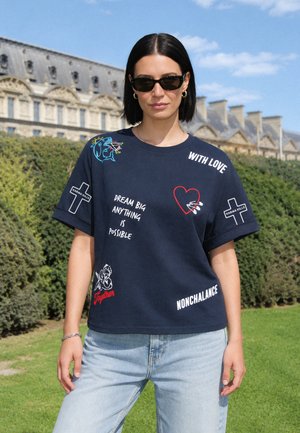 Femme aux cheveux courts noirs portant des lunettes de soleil noires, un tee-shirt graphique bleu marine avec du texte et des symboles, et un jean bleu clair en extérieur près de buissons taillés.