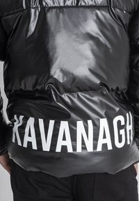 Giacca a vento nera e lucida con un prominente testo bianco "KAVANAGH" sul retro, con una superficie liscia e orlo elastico.