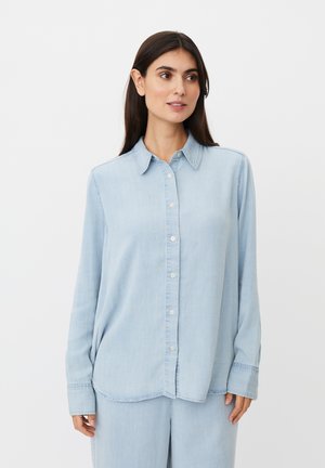 Vrouw met lang donker haar, een lichtblauw denimoverhemd met knopen en bijpassende broek, kijkt iets naar de zijkant.