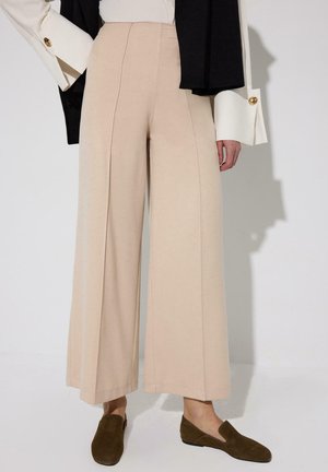 Pantalon beige à jambes larges associé à des chaussures plates vert olive, blouse blanche avec manches longues oversize, et veste noire portée sur l'épaule.