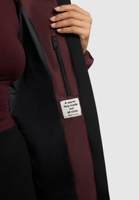 Chaqueta de concha negra con forro burdeos. Cuenta con un bolsillo con cremallera y una etiqueta que dice "Un abrazo cálido hecho de tela." Textura suave.