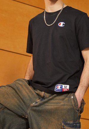 Junger Mann trägt ein schwarzes Champion-T-Shirt mit Logopatches und olivgrünen Cargohosen, lehnt mit den Händen in den Taschen an einem hölzernen Hintergrund.