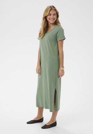 Femeie într-o rochie midi verde salvie, cu mâneci scurte și decolteu în V, cu o despicătură laterală, purtând balerini negri și bijuterii aurii, zâmbitoare și uitându-se într-o parte.