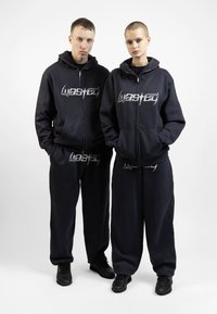 Dark navy hoodies and sweatpants with the word "Wasted" in silver lettering. Both garments feature a relaxed fit and unzippered pockets.