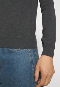 Pull gris foncé à manches longues avec poignets et ourlet côtelés, associé à un jean bleu en denim, montrant un gros plan du torse et de la main droite.