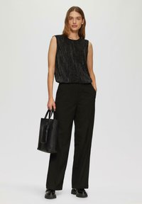 Haut sans manches noir avec des plis verticaux texturés, associé à un pantalon noir taille haute. Tenant un petit sac fourre-tout noir. Chaussures noires.