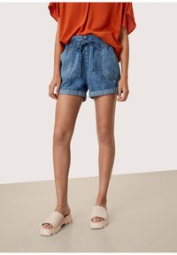 Denim shorts with an elastic waistband and drawstring, featuring two front pockets and rolled hems. Worn with beige platform slide sandals.