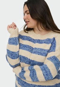 Beige trui met blauwe strepen, voorzien van een ronde hals en geribde manchetten. Gebreide textuur met verschillende tinten en patronen.