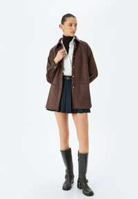 Chaqueta de ante sintético marrón con cuello, botones en la parte frontal y bolsillos laterales, llevada sobre una camisa blanca y una falda plisada oscura, combinada con botas hasta la rodilla.