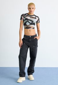 Cropped, sheer black top with abstract colorful pattern; high-waisted, dark cargo pants with pockets and hardware details; neutral sneakers.