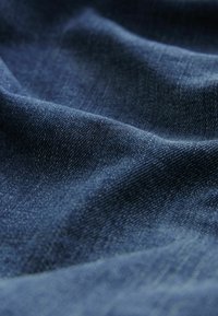 Donkerblauwe denim stof met een gestructureerd oppervlak, dat plooien en kreukels laat zien en het weefpatroon en een lichte glans benadrukt.