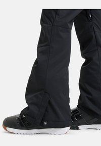 Zwarte sneeuwbroeken met een getailleerde pasvorm en een gladde, gestructureerde afwerking. Glanzende zwarte sneeuwlaarzen met witte zolen en zwarte details worden gedragen.