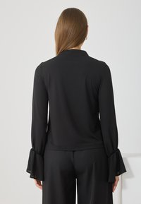 Blusa nera a maniche lunghe con texture liscia, caratterizzata da polsini a campana e vestibilità sartoriale, vista posteriore che mette in evidenza il design delle maniche e l'orlo.
