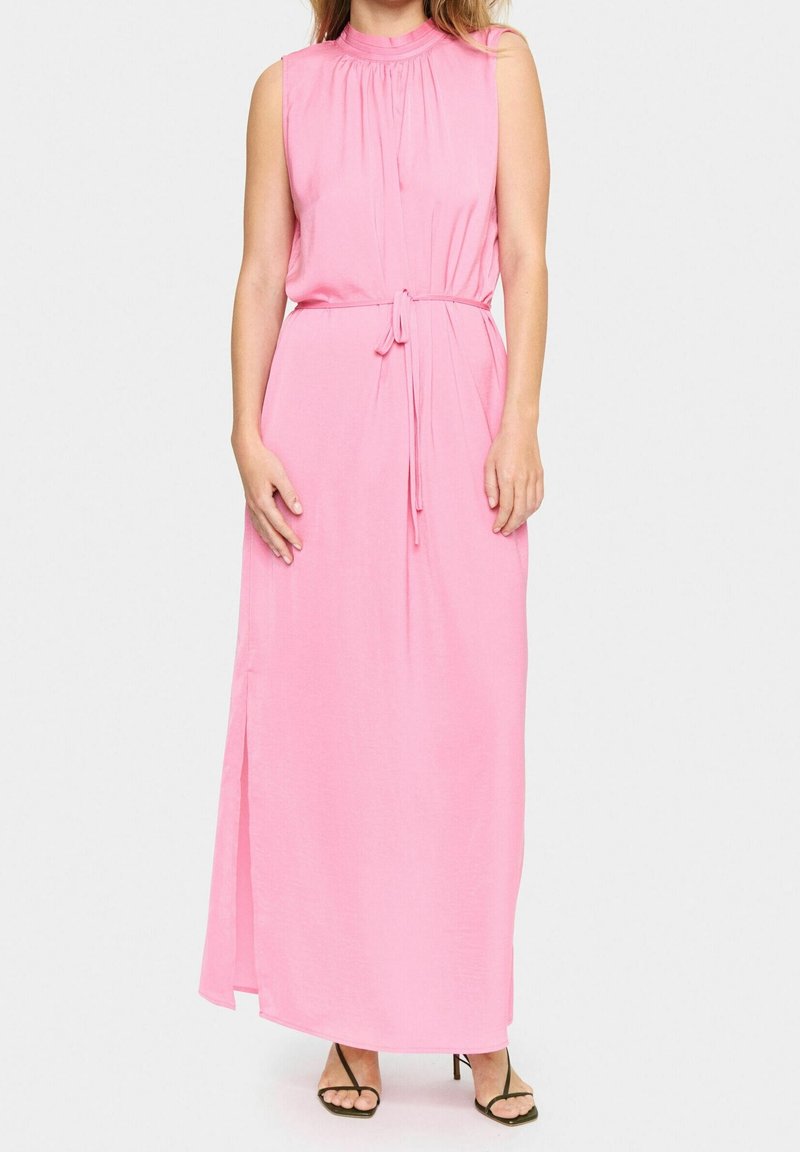 Femme portant une robe maxi rose sans manches avec un col froncé, une ceinture nouée à la taille, des fentes latérales et des talons ouverts noirs, debout sur fond blanc.