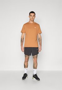 Oransje bomull T-skjorte med korte ermer og en liten logo, sammen med svarte shorts. Svarte joggesko og hvite sokker fullfører antrekket.