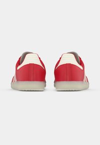 Par de ténis vermelhos com detalhes brancos no calcanhar e solas translúcidas, vista de trás sobre um fundo branco simples.