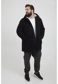 Svart parkasjakke med hette, med en krage foret med pels, frontlommer og snor i livet. Stilt med grå bukser og svarte joggesko.