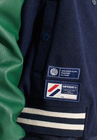 Marineblauw jasje met groene leren mouwen, wollen lijf, gestreepte geribde zoom en meerdere merkpatches aan de linkerkant.