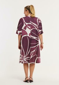 Blomstret kjole i dyb lilla og hvid. Korte ærmer, løs pasform, midtlang. Har store blomsterprint og en lige skæring bagpå.