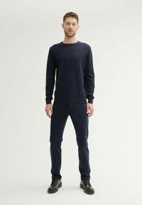 Pull à manches longues bleu marine en tricot doux, associé à un pantalon bleu marine et des chaussures noires à lacets. Design minimaliste, sans motifs ni logos visibles.