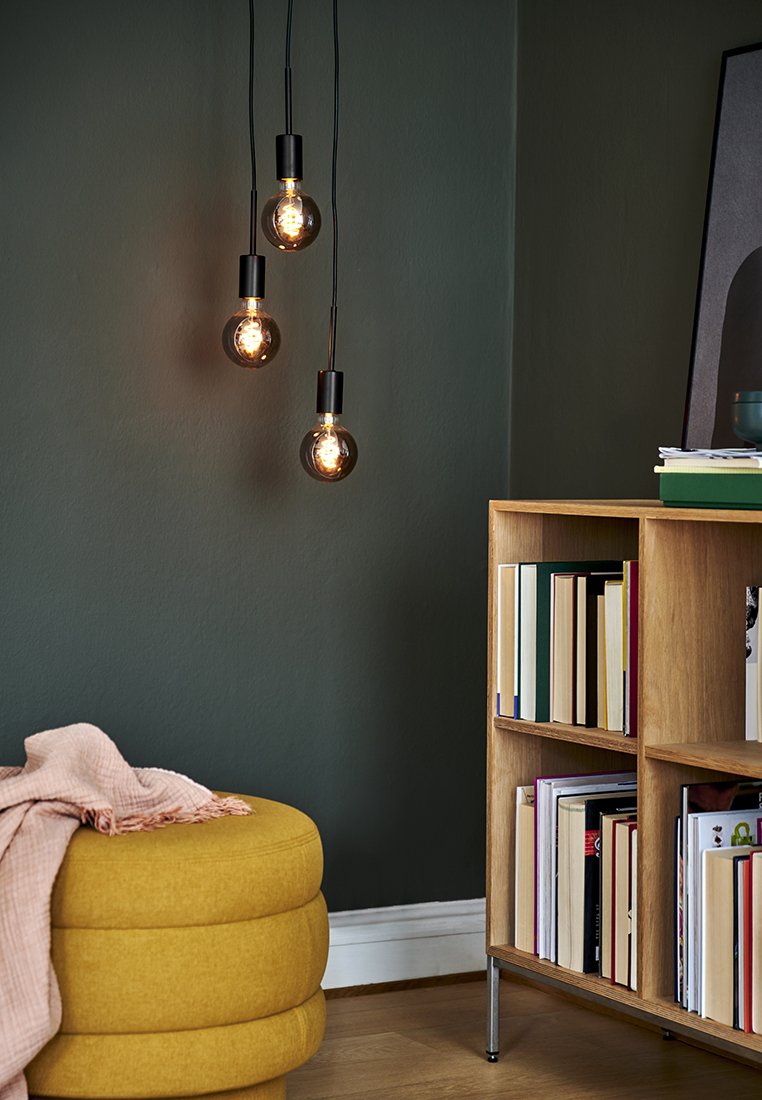 Trzy zapalone wiszące żarówki w pobliżu drewnianej regału wypełnionego książkami oraz musztardowo żółtego pufy z różowym kocem narzuconym na niego.