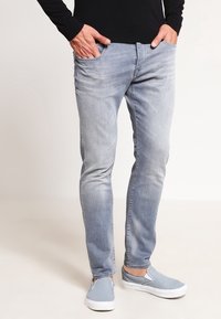 Jeans ajustados de color azul claro con un lavado desgastado y un sutil efecto de arrugas. Presenta bolsillos frontales y una clásica cremallera. Combinados con zapatillas informales.