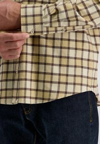 Geel en bruin geruit shirt met knoopmanchetten. De stof is geweven en toont een ruitpatroon, gestopt in een donkere spijkerbroek.