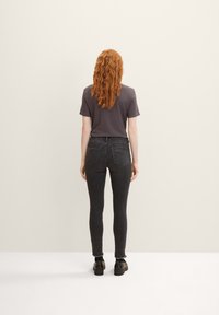 Grijze t-shirt, zwarte skinny jeans met een rafelrand, en zwarte schoenen. De persoon staat met de rug naar de kijker tegen een neutrale achtergrond.