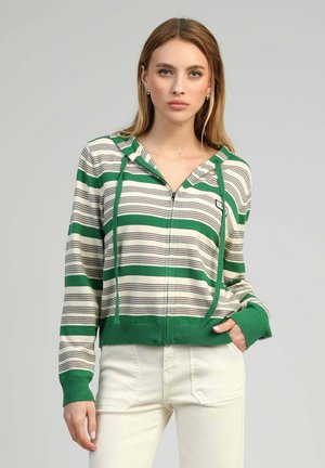 Jeune femme portant un sweat à capuche zippé rayé vert et blanc et un pantalon blanc, debout devant un fond gris clair uni.