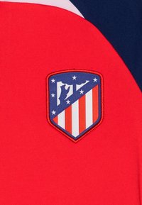 Tessuto rosso con un ricamo del logo del club calcistico Atlético Madrid, caratterizzato da strisce rosse e bianche e una sezione superiore blu con un orso e un albero.