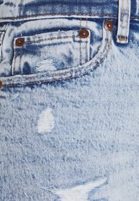 Tela de mezclilla azul clara con detalles desgastados, un pequeño bolsillo frontal y acentos de hardware en color cobre. Texturizada con bordes deshilachados.