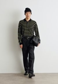 Polo cu dungi negre și măslinii, cu mânecă lungă, pantaloni negri și o căciulă neagră. Ținând o geantă mare neagră; stând pe un fundal simplu.